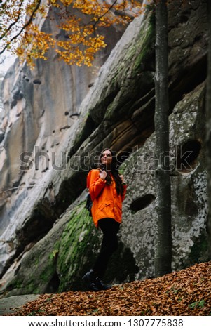 Young woman wearing warm orange sweater standing on a fallen autumn leaves in the autumn forest. Misty landscape with mossy rocks. Cute smiley woman in the nature. Autumn forest hiking