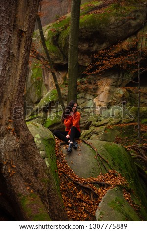Young woman wearing warm orange sweater sitting on the rock in the autumn forest. Misty landscape with mossy rocks. Cute smiley woman in the nature. Autumn forest hiking