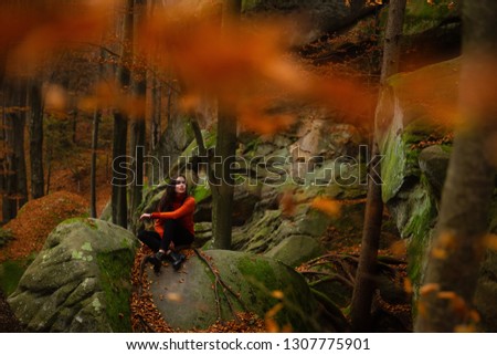 Young woman wearing warm orange sweater sitting on the rock in the autumn forest. Misty landscape with mossy rocks. Cute smiley woman in the nature. Autumn forest hiking