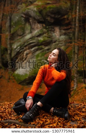 Young woman wearing warm orange sweater sitting on a fallen autumn leaves in the autumn forest. Misty landscape with mossy rocks. Cute smiley woman in the nature. Autumn forest hiking