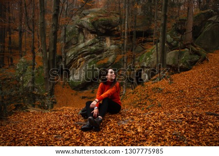 Young woman wearing warm orange sweater sitting on a fallen autumn leaves in the autumn forest. Misty landscape with mossy rocks. Cute smiley woman in the nature. Autumn forest hiking