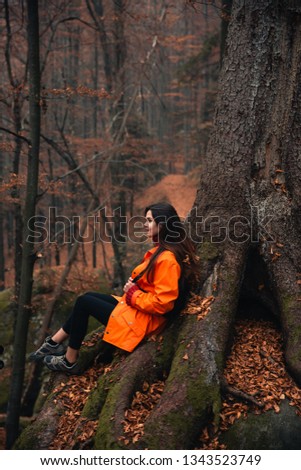 Young woman wearing warm orange jacket sitting on the rock in the autumn forest. Misty landscape with mossy rocks. Cute smiley woman in the nature. Autumn forest hiking