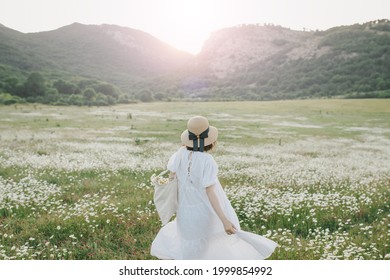 Young Woman Wearing White Dress Holding Cotton Bag With Flowers On Chamomile Field. Cottagecore Aesthetic.