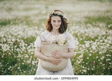 Young Woman Wearing White Dress Holding Straw Basket With Flowers On Chamomile Field. Cottagecore Aesthetic.