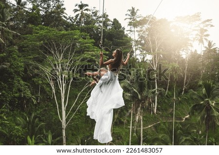 Young woman wearing white dress swinging on rope swings with beautiful view on rice terraces and palm trees in the Bali Island