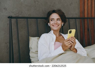 Young woman wearing white shirt pajama she lying in bed use mobile cell phone listen music in earphones rest relax spend time in bedroom lounge home in own room hotel wake up dream in good mood day. - Shutterstock ID 2206212525
