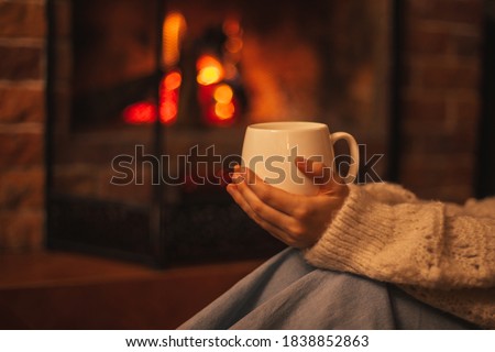 Young woman wearing white woolen knitted sweater enjoying hot tea near fireplace in a cozy living room in the evening.