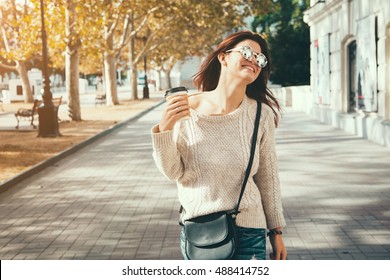 Young Woman Wearing Woolen Sweater Walking In The Autumn City Street And Drinking Take Away Coffee In Paper Cup. Breakfast On The Go.