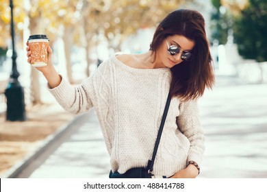 Young woman wearing woolen sweater walking in the autumn city street and drinking take away coffee in paper cup. Breakfast on the go.