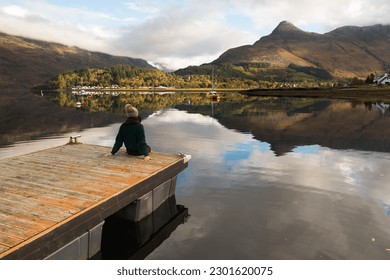 Young woman wearing a woolly hat sitting on the edge of a pier overlooking the mirror-like water of the Loch Leven with mountains on the background and boats floating on the water in Glen Coe