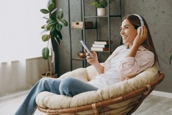 Young Woman Wears Casual Clothes Sits In Armchair Listen To Music In Headphones Use Mobile Cell Phone Stay At Home Hotel Flat Rest Spend Free Spare Time In Living Room Indoor. Lifestyle Lounge Concept