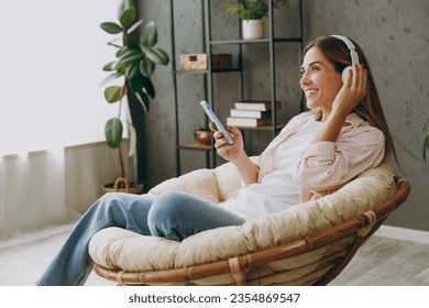 Young woman wears casual clothes sits in armchair listen to music in headphones use mobile cell phone stay at home hotel flat rest spend free spare time in living room indoor. Lifestyle lounge concept