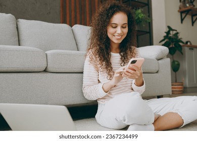 Young IT woman wears casual clothes sits near grey sofa couch use mobile cell phone laptop pc computer stay at home hotel flat rest relax spend free spare time in living room indoor. Lounge concept - Shutterstock ID 2367234499
