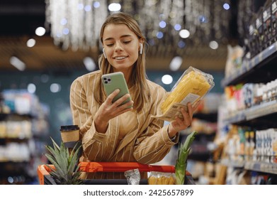 Young woman wears casual clothes earphones listen music use mobile cell phone hold pasta shopping at supermaket store grocery shop buy with cart choose products in hypermarket. Purchasing food concept