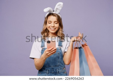Young woman wears casual clothes bunny rabbit ears hold paper package bag after shopping use mobile cell phone isolated on plain light pastel purple background studio Happy Easter sale buy day concept