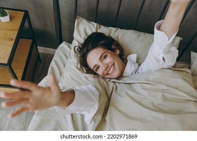 Young woman wears casual white shirt she lying in bed rest relax spend time in bedroom lounge home in own room hotel wake up stretch hands dream be lost in reverie good mood day. Lazy morning concept