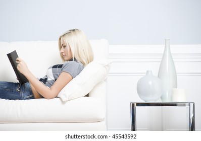 Young Woman Wears A Checkered Top And Blue Jeans, Lying On White Sofa And Reading Book. Hoizontal Shot.