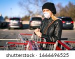 A young woman wears an N95 respirator while leaning on a shopping cart outside of a grocery store. The woman is protecting herself from coronavirus and other airborne particles and diseases. 