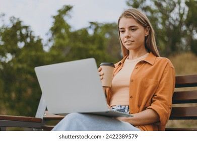 Young IT woman wears orange shirt casual clothes hold use work on laptop pc computer drink coffee sit on bench walk rest relax in spring city park outdoors on nature. Urban lifestyle leisure concept - Powered by Shutterstock