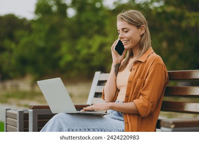 Young woman wears orange shirt casual clothes hold use work on laptop pc computer talk on mobile cell phone sit on bench walk rest relax in spring city park outdoors on nature. Urban lifestyle concept - Powered by Shutterstock