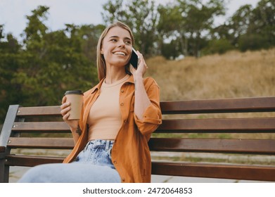 Young woman wears orange shirt casual clothes talk speak mobile cell phone look aside drink coffee sit on bench walk rest relax in spring green city park outdoors on nature. Urban lifestyle concept - Powered by Shutterstock