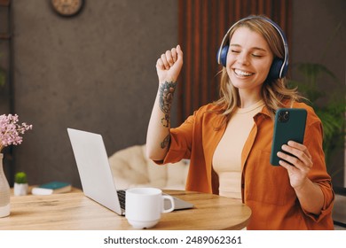 Young IT woman wears orange shirt headphones hold mobile cell phone use laptop pc computer listen music sit alone at table in coffee shop cafe indoor work or study. Freelance office business concept