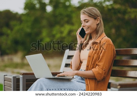 Young woman wears orange shirt casual clothes hold use work on laptop pc computer talk on mobile cell phone sit on bench walk rest relax in spring city park outdoors on nature. Urban lifestyle concept