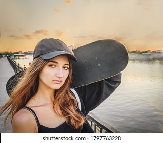 A young woman wears a T-shirt with straps and a black hat, she holds a skateboard on her shoulder and looks at the camera. Loneliness in town. The river and the painterly sunset sky in the background.