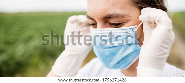 Young woman wears a\
virus protection mask and wears surgical sterile gloves on her\
hands. Girl standing in the green field smiling and putting on a\
protective face mask.