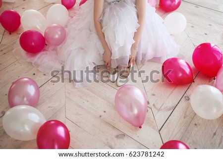 Young woman in wedding dress in luxury interior with a mass of pink and white balloons, sitting on the floor. Hold in hands her white shoes.