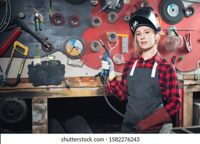 Young woman welder smiles and holds protective mask in hand on background industrial workplace or garage. Concept small business craft.