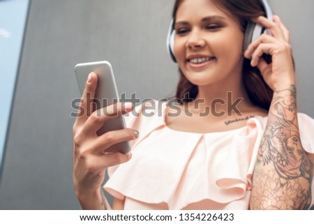 Young woman wering headphones standing isolated on grey wall on the city street listening to favorite song holding smartphone browsing playlist close-up smiling happy