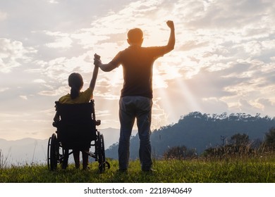 Young Woman In Wheelchair Raised Hands With His Care Helper In Sunset.Silhouette