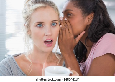 Young woman whispering secret to her shocked friend at home