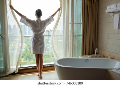 Young Woman In White Bathrobe Opening Window In Luxury Bathroom With City View