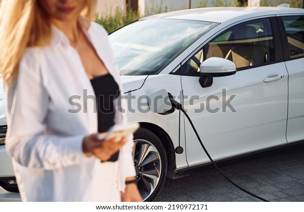 Young woman in white clothes is with her\
electric car at daytime.