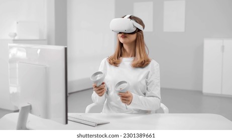 Young woman in white clothes and vr headset sitting at desk in white room. Female work in augmented reality goggles using joysticks - Powered by Shutterstock