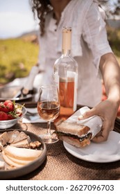 Mujer joven en vestido blanco que sostiene el bocadillo con la tabla de la comida campestre, la placa del vino, y el vino rosado