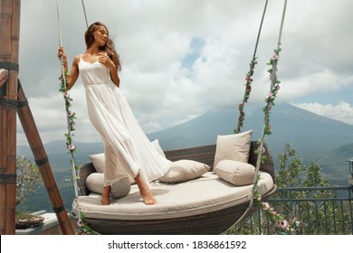 Young Woman In White Dress Posing On Swing Against Volcano Agung In Bali, Indonesia. Beautiful Girl Standing On Hanging Chair With Flowers On Mountain Background.  