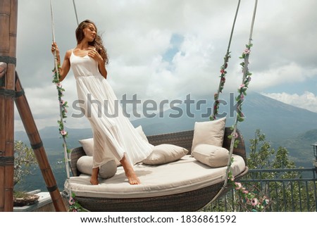 Young Woman In White Dress Posing On Swing Against Volcano Agung In Bali, Indonesia. Beautiful Girl Standing On Hanging Chair With Flowers On Mountain Background.  