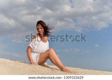 A young woman in a white dress sitting barefoot on the sandy beach in the hot summer.