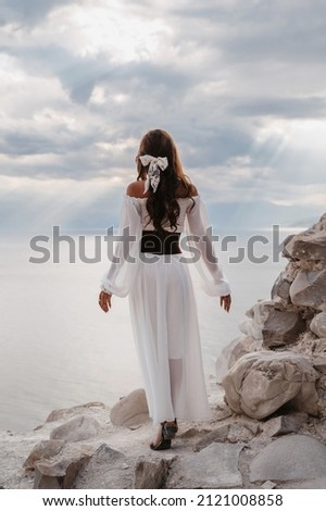 A young woman in a white dress stands barefoot on a cliff face in full height. The dress flutters in the wind. A sacred glow breaks through the clouds. The girl looks like an angel