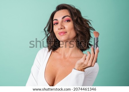 Young woman in a white evening dress on a light green background with bright pink makeup holding a blush brush
