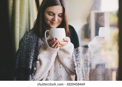 Young woman in white knitted sweater drinking tea near window in a cozy house. - Shutterstock ID 1820458469