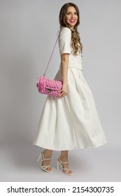 Young Woman In A White Maxi Dress And High Heels Is Doing A Turn While Holding A Pink Braided Purse Over Her Shoulder. Full Length Studio Shot.