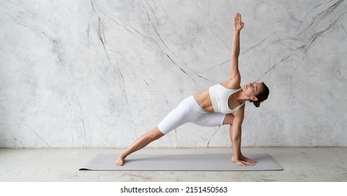 Young woman in white outfit practicing yoga bikram triangle, trikonasana in trendy minimalist studio with copy space on the textured wall