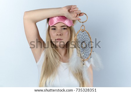 young woman in white shirt going sleep in a sleep mask and dream catcher in hand. Portrait on white background, expressive look at the camera.