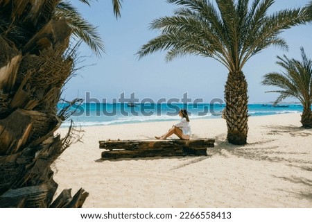 Young woman in white shirt, shorts and sunglasses sitting on a wooden bench on a sunny sandy beach surrounded by palm trees and looking at the turquoise ocean in Santa Maria, Sal island, Cape Verde.