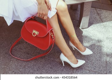 Young Woman In White Summer Classic Leather High Heel Shoes With Bright Red Leather Handbag. Girl In A White Dress With Glitter Tattoo On Her Leg Resting On The Summer Terrace. Handbag Fashion Look