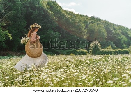 A young woman in a white sundress, a wreath of daisies with a large wicker bag on her shoulder is walking through a field of daisies, against the background of the forest, the sunset light